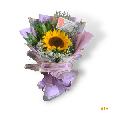Bia arrangement on a plain background: Cheerful sunflower arrangement with white fillers and fresh greenery, wrapped in soft lavender and pink tones.