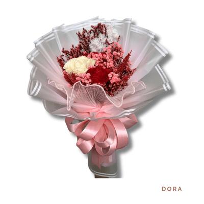 Dora arrangement on a plain background: Delicate dried bouquet of cream and red blooms with soft foliage, wrapped in sheer pink and finished with a voluminous pink ribbon.