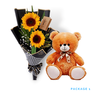Two sunflowers with greenery and baby's breath in black wrap beside a seated light-brown teddy bear with a ribbon bow, on white