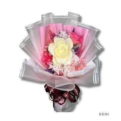 A dried bouquet of a cream peony, pink wood roses, blush hydrangea and purple grasses in layered pink and white wrap, tied with a dark ribbon, on white